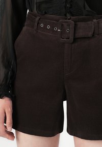 Shorts en velours côtelé marron foncé avec une ceinture en tissu assortie et des œillets en métal argenté, portés avec un chemisier noir en tissu léger à manches longues.