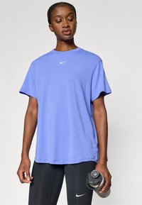 Μωβ αθλητική κοντομάνικη μπλούζα με στρογγυλή λαιμόκοψη, με διακριτικό λογότυπο Nike στο στήθος. Φορεμένη με μαύρα κολάν και κρατώντας ένα μπουκάλι νερό.