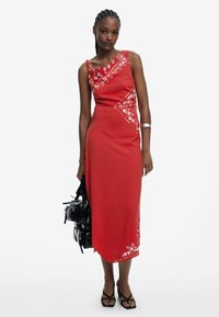 Kleid punah tanpa lengan berwarna merah dengan bordir bunga putih, celah samping, dan siluet lurus. Dipadukan dengan tas hitam dan sandal bertumit tinggi.