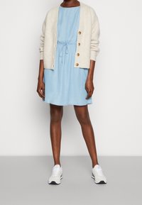 Svetle modré džínové šaty s širokým pasem na stahovací šňůrku, doplněné krémovým žebrovaným kardiganem se třemi velkými knoflíky. Na nohou bílé tenisky.