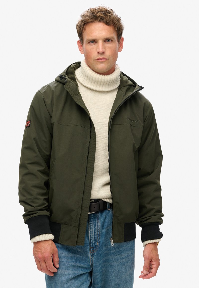 Olivengrøn jakke med hætte, lynlås-lukning og ribbede manchetter. Båret over en cremefarvet turtleneck-trøje og blå jeans. Simpelt og funktionelt design.