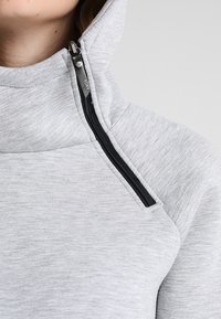 Sweat-shirt gris clair avec un col montant, doté d'une ouverture zippée, d'un tissu texturé et d'un élégant accent de fermeture éclair noire.