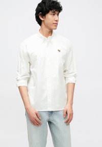 Λευκό πουκάμισο με κουμπιά από βαμβάκι, με μικρό κεντητό σχέδιο ενός ελαφιού στην αριστερή πλευρά του στήθους. Φορεμένο με ανοιχτόχρωμο μπλε τζιν.