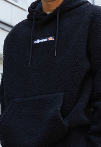 Felpa nera con cappuccio in pile, tasca a marsupio, cappuccio con coulisse regolabile e logo bianco sul petto con accenti rossi e gialli.