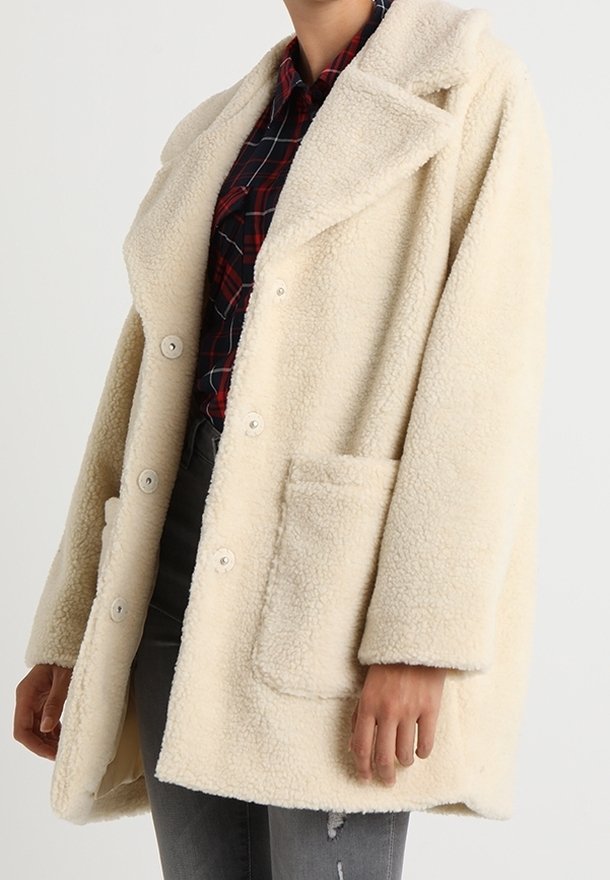 Persona che indossa un cappotto sherpa crema oversize sopra una camicia a scacchi rossa e blu navale con jeans scuri strappati, mani rilassate ai lati.