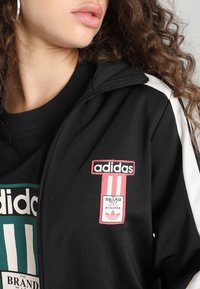 Svart zip-up jacka med vita detaljer, som har en broderad röd Adidas-logga och en rektangulär patch med text. Material av bomullsblandning.