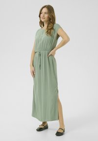 Long, flowing dress in light green fabric, featuring short sleeves, elastic waistband, side slits, and paired with black flat shoes.