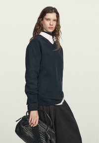 Marineblauwe v-hals trui gemaakt van zachte bre Stoffen, met lange mouwen en een ontspannen pasvorm, gecombineerd met een zwarte geweven tas en over een wit shirt gedrapeerd.