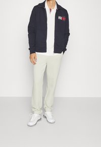 Giacca zip-up blu navy con logo e grafica dell'anno, abbinata a una polo bianca e pantaloni tuta grigi chiari, completata da sneaker bianche.