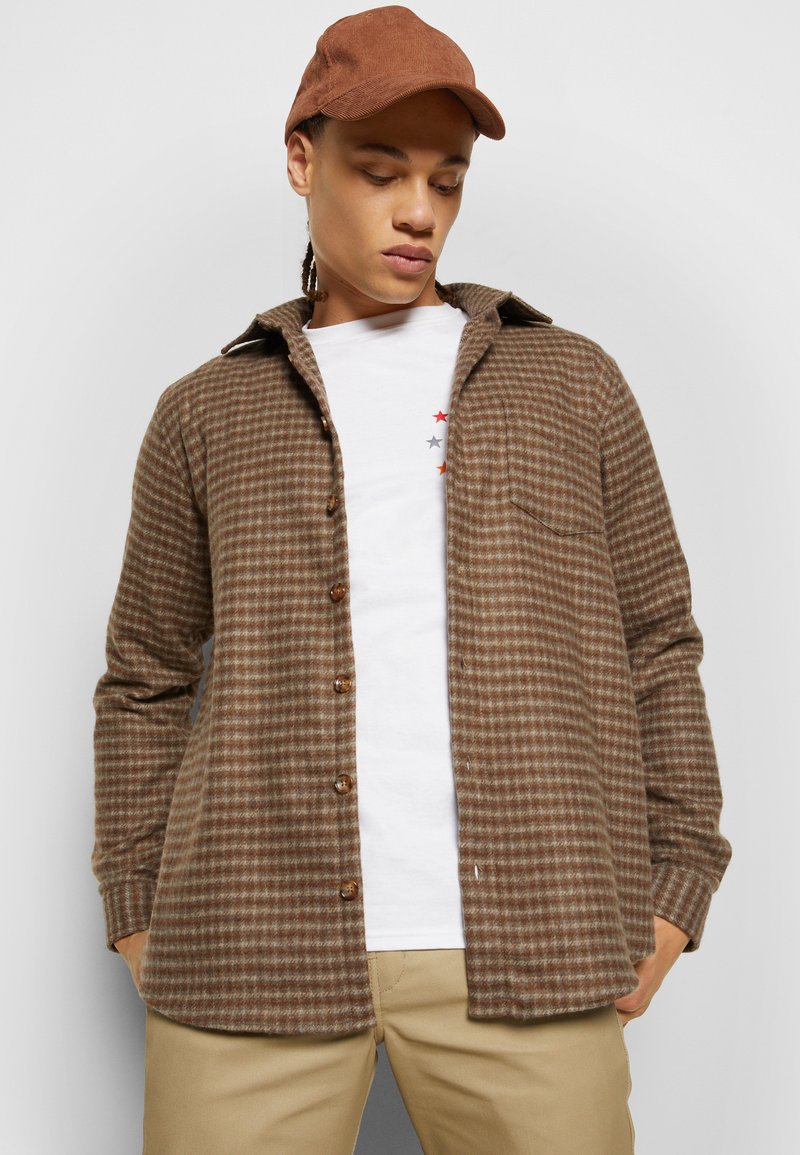 Chemise à carreaux marron avec un devant à boutons, un col et une poche sur la poitrine. Portée sur un t-shirt blanc avec des graphismes d'étoiles colorées.