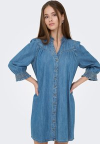 Νεαρή γυναίκα που φοράει μπλε φόρεμα από ντένιμ με κουμπιά μπροστά, φουσκωτά μανίκια και βολάν στις μανσέτες, στέκεται με τα χέρια στη μέση μπροστά από ένα μονόχρωμο φόντο.