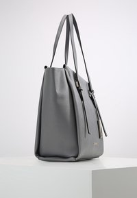 Sac fourre-tout en cuir gris avec surface texturée, deux fines bandoulières et détail étiquette en métal, posé sur un socle blanc sur un fond uni.