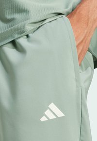 Světlé zelené sportovní šortky vyrobené z hladké látky, s vypouklou texturou, boční kapsou a bílým diagonálním logem se třemi pruhy.