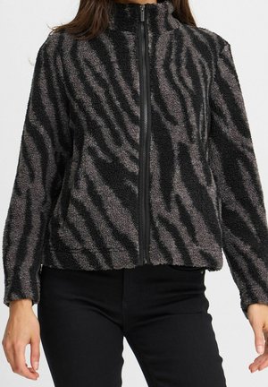 Veste en polaire noire et grise à motif zèbre avec fermeture éclair à l'avant, portée avec un pantalon noir.