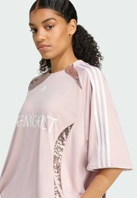 Camiseta deportiva rosa con franjas blancas en las mangas, acentos brillantes en los hombros y texto "TEAMGEIST" en el frente. Corte corto y holgado.