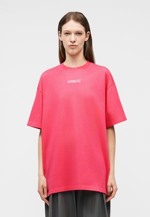 Νεαρή γυναίκα που φοράει ένα υπερμεγέθες, φωτεινό ροζ T-shirt με το κείμενο "STIEGLITZ", στέκεται μπροστά σε ένα απλό λευκό φόντο.