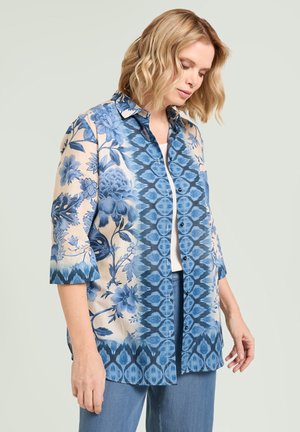 Femme aux cheveux blonds ondulés portant une chemise boutonnée à motifs floraux bleu et beige et un jean bleu, regardant vers le bas.