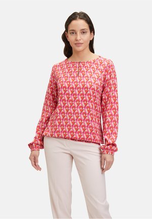 Langärmlige Bluse in leuchtendem Pink mit geometrischem Druck, mit runder Ausschnitt und gerafften Manschetten, kombiniert mit hellen Hosen.