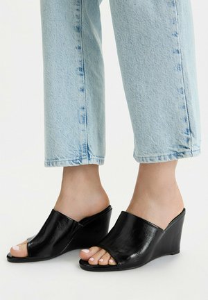 Pies con vaqueros azul claro y sandalias de cuña negras de cuero con punta abierta sobre fondo blanco.
