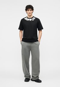 Ung mand med krøllet hår iført en sort t-shirt med teksten "BARRON" og løse grå bukser, stående med hænderne i lommerne.