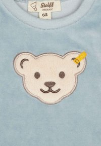 Lichtblauwe sweatshirt met een gestructureerde berenface patch in roomwit en bruin, met een gele label op het oor van de beer. Gemaakt van biologisch materiaal.