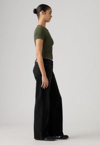 Πράσινο εφαρμοστό τοπ με κοντά μανίκια συνδυασμένο με μαύρο τζιν παντελόνι σε φαρδιά γραμμή και ψηλή μέση. Το σύνολο έχει λεία υφή και casual σχεδιασμό.