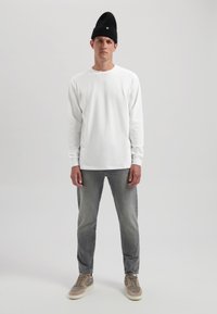 Fehér hosszú ujjú ing, szürke slim-fit farmernadrág és szürke sportcipő. Fekete kötött sapka kis zöld díszítéssel, egy sima háttér előtt áll.