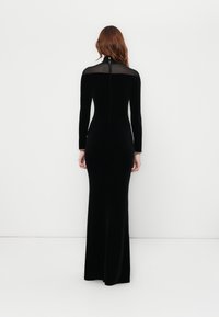 Black velvet long-sleeve gown with sheer upper panel, fitted silhouette, and zipper closure at the back. Gold chain accent at wrist.