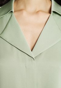 Ljusgrön blus med spetsig krage, slät yta och subtila sömdetaljer, med en V-formad halsringning.