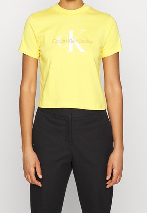 Osoba ubrana w żółty krótki top z białym logo "Calvin Klein Jeans" i czarne spodnie z wysokim stanem, widoczny tors.