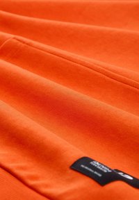 Tela de algodón naranja con una textura suave, que presenta costuras cosidas y una pequeña etiqueta negra con texto blanco en el lado derecho.