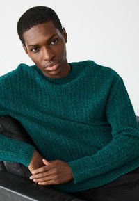 Sinine tekstureeritud sviiter, mis on valmistatud kudumiskangast, ümarate kaelustega ja soonikute mansettidega, koos mustade riietega. Pehme ja soe välimus.