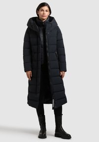 Kvinde stående iført en lang sort dunjakke med hætte, sorte leggings og sorte læderankelstøvler på en ensartet lys baggrund.