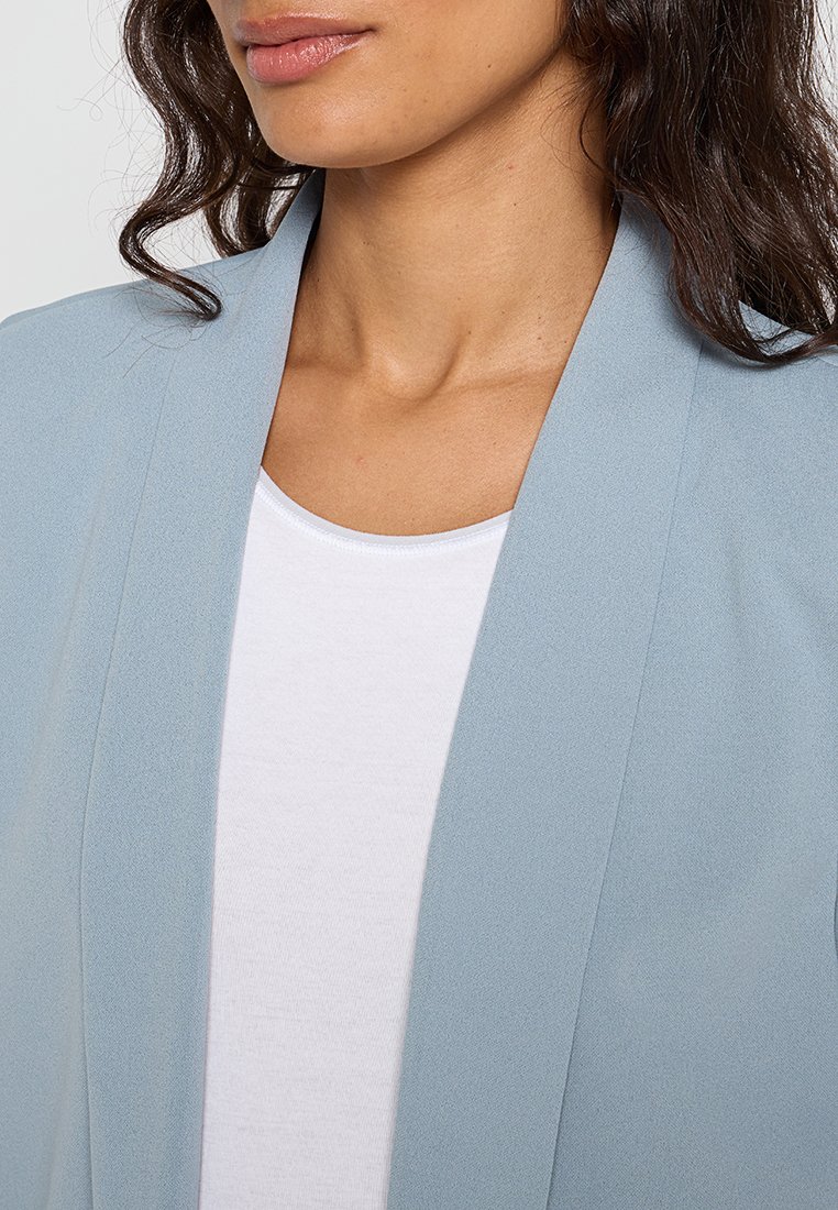 Blazer bleu clair en tissu doux avec une coupe décontractée, à l'avant ouvert et sans fermetures visibles, superposé sur un haut blanc.