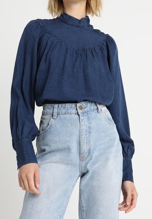 Frau trägt eine marineblaue Bluse mit hohem Kragen und langen Puffärmeln, kombiniert mit hochtaillierten hellblauen Jeans.