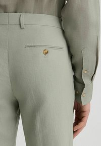 Hellgrüne Leinenhosen mit einer Gesäßtasche und einem goldenen Knopf. Der Stoff hat eine strukturierte Oberfläche, und der Bund ist glatt.