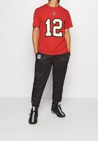 Czerwona koszulka z krótkim rękawem z czarnym numerem "12" i logo, zestawiona z czarnymi spodniami z logiem NFL oraz czarnymi trampkami z srebrnymi akcentami.