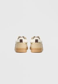Vue arrière de baskets beiges avec une tige crème, des accents dorés et une finition texturée. Elles disposent d'une languette marron au talon et d'une semelle en caoutchouc.
