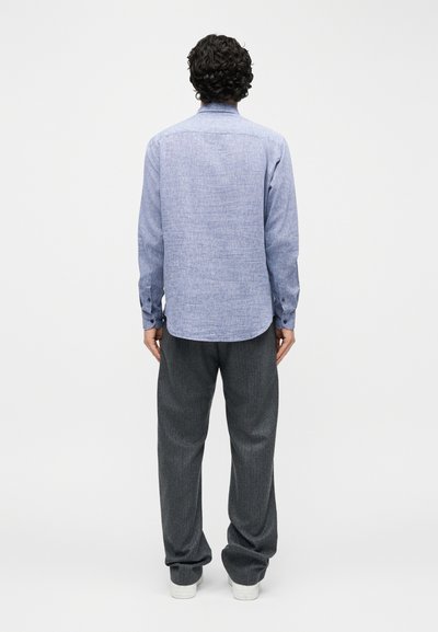 Άνδρας με σγουρά σκούρα μαλλιά γυρισμένος με την πλάτη, φορώντας ανοιχτό μπλε μακρυμάνικο πουκάμισο, σκούρο γκρι παντελόνι και λευκά παπούτσια, σε απλό φόντο.