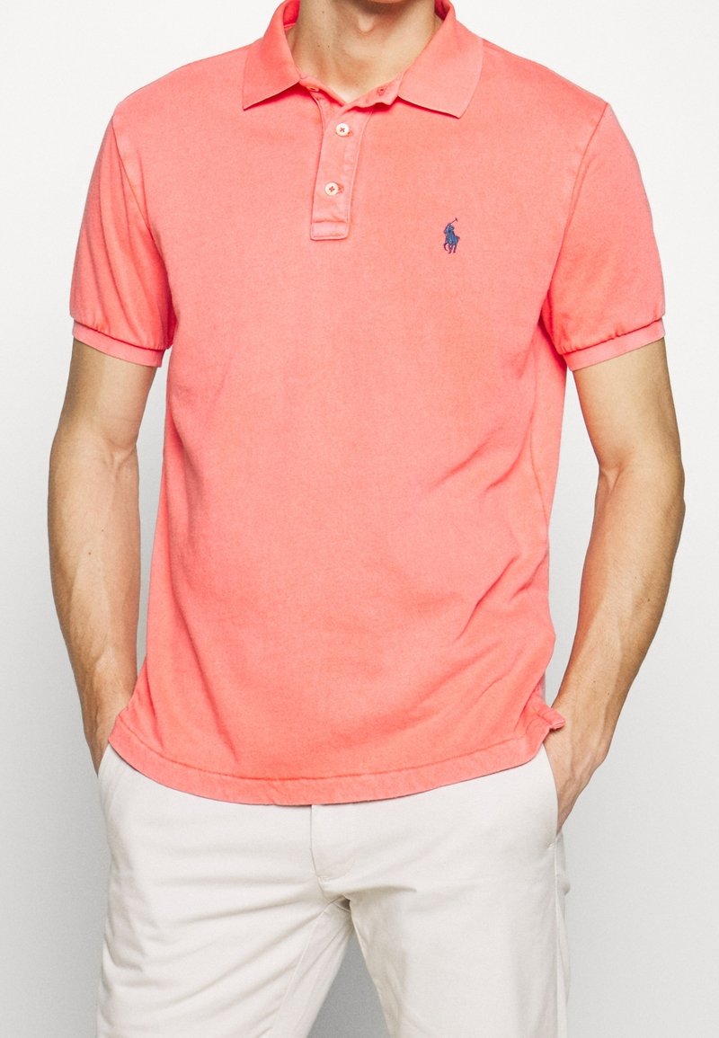 Homme portant un polo corail vif à manches courtes avec col et un pantalon blanc, mains dans les poches, avec un petit logo brodé sur la poitrine.