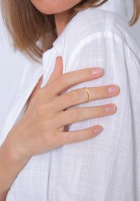 Kuldne metallrings sõrmel, mis resting tekstureeritud valgel linases särgil. Kanga peal on kerge ruuduline muster koos pehmete voldidega.