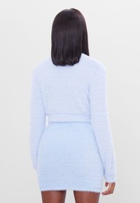 Donna con capelli scuri e lisci, che indossa un maglione lungo a maniche lunghe di un blu chiaro morbido e una mini gonna abbinata, in piedi contro uno sfondo chiaro e semplice.