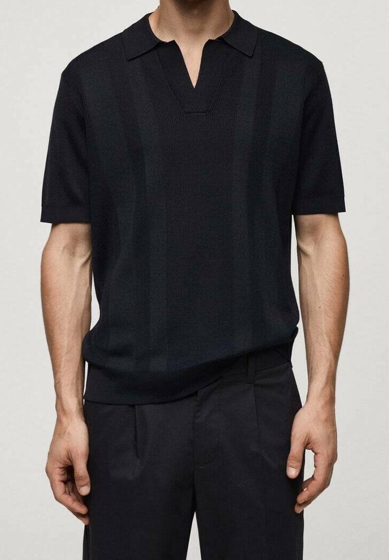 Homme portant un polo tricoté noir à manches courtes avec un col en V, associé à un pantalon noir, sur un fond uni.