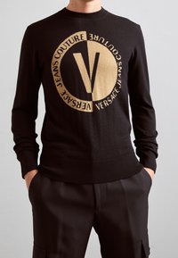 Pull en maille noire avec encolure ronde, présentant un grand logo beige et un texte. Poignets et ourlet côtelés, avec une texture lisse.