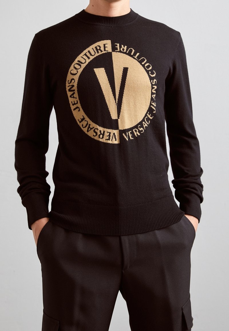 Pull en maille noire avec encolure ronde, présentant un grand logo beige et un texte. Poignets et ourlet côtelés, avec une texture lisse.