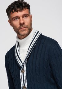 Cardigan blu navy in maglia a treccia con collo a scialle, dettagli a righe bianche e bottoni in legno, indossato sopra un maglione bianco a collo alto.