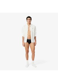 Witte button-up blouse, zwarte Lacoste boxershorts en witte sokken. De blouse heeft een borstzak met een groen logo. Glad materiaal, strakke pasvorm.