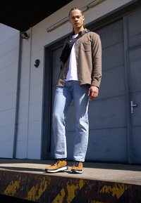 Hnědě kostkovaná košile přes bílý tričko, světlé modré džíny a hnědé vysoké boty s černými akcenty, stojící na nakládací rampě.