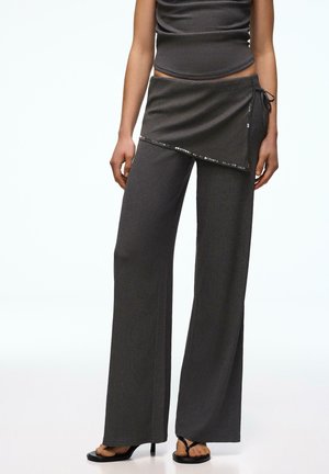 Nő sötétszürke bordázott, bőszárú nadrágot visel, amelyhez egy oldalt megkötős, flitteres szegéllyel díszített csatolt wrap szoknya tartozik, fekete magas sarkú szandállal párosítva.