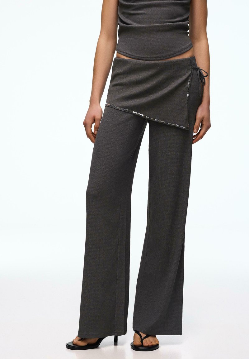 Femeie purtând pantaloni largi cu talie înaltă, gri închis, cu dungi, având o fustă înfășurată atașată, cu un nod lateral și margine cu paiete, asortați cu sandale negre cu toc.